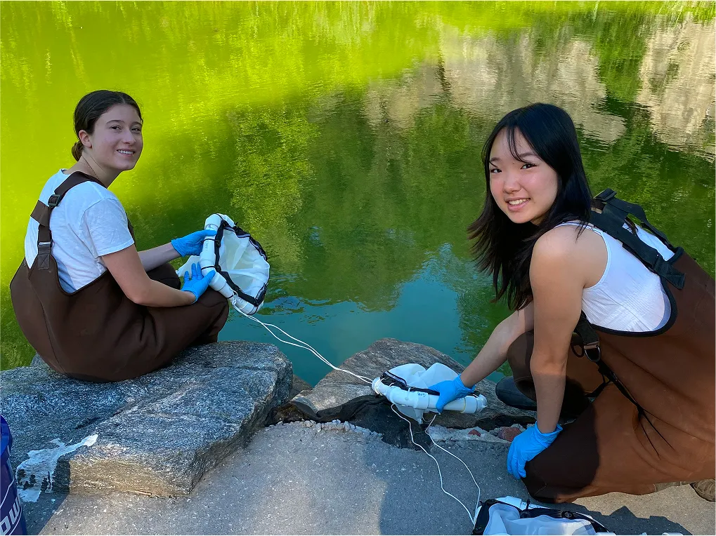Two young women in waders putting nets in a green pond.
