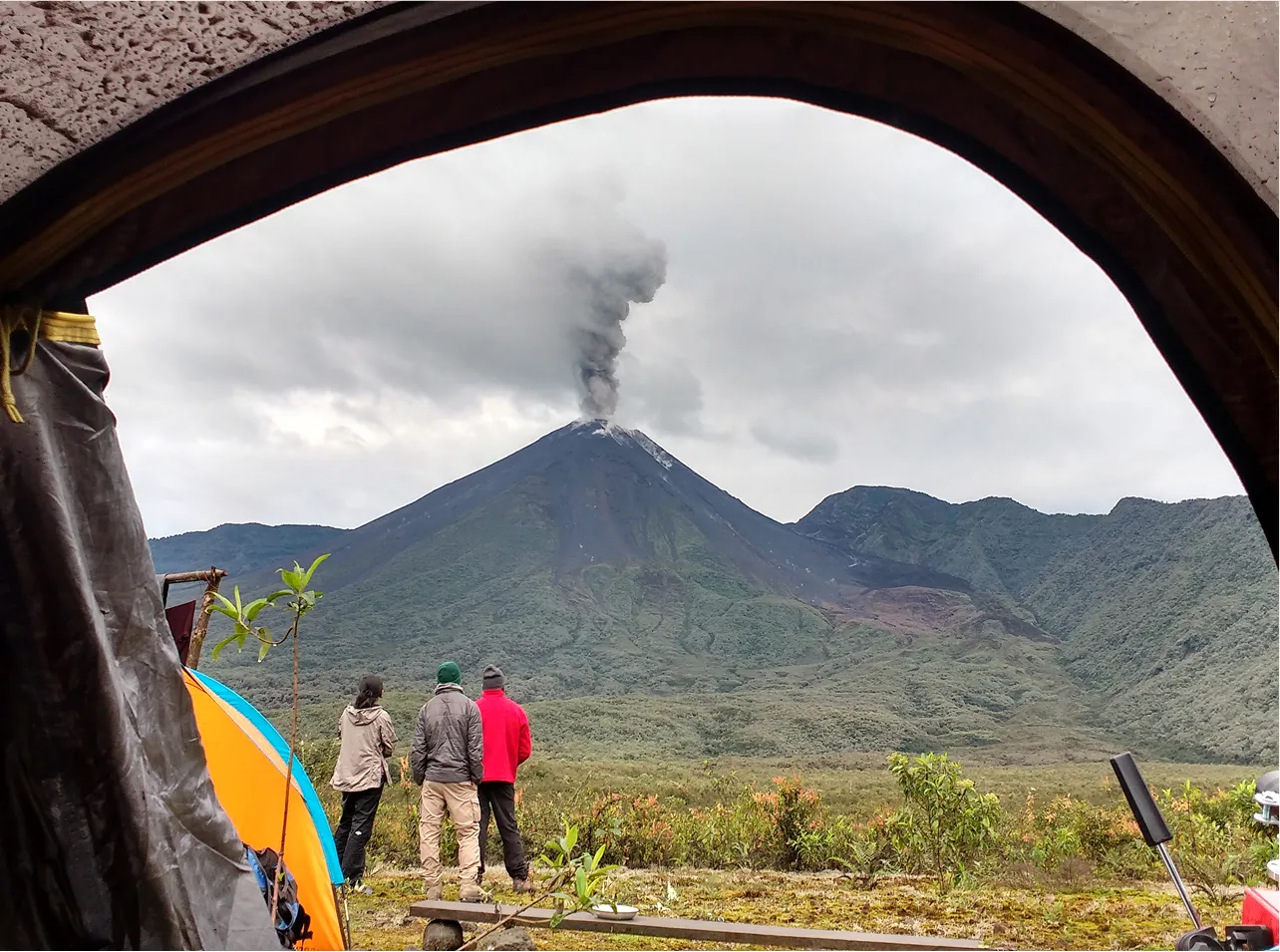 Three people seen through an archway looking at a smoking volcano.