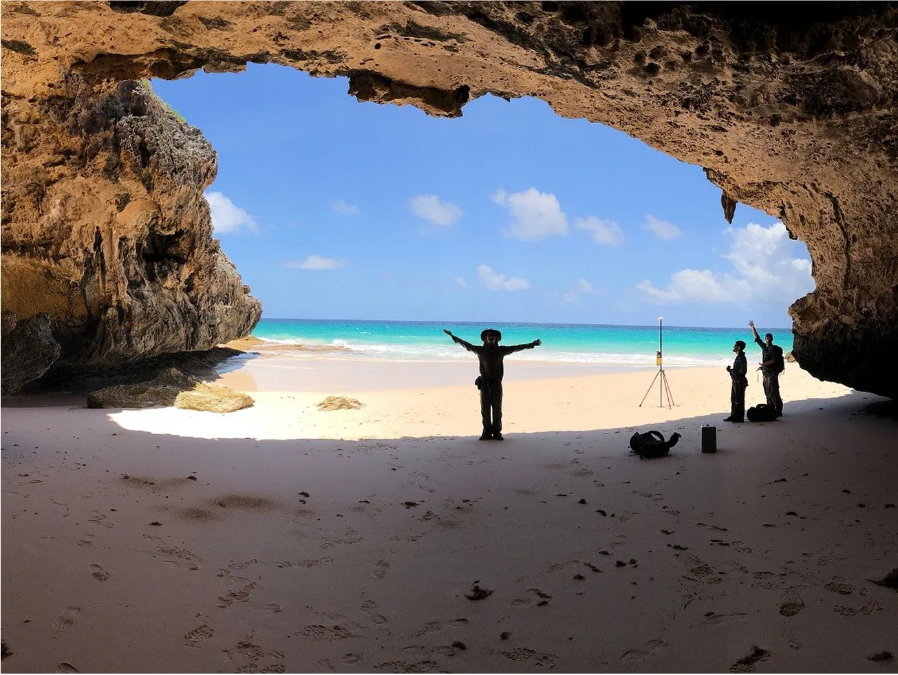 Person with outstretched arms under a rock formation in front of a blue ocean and sky view
